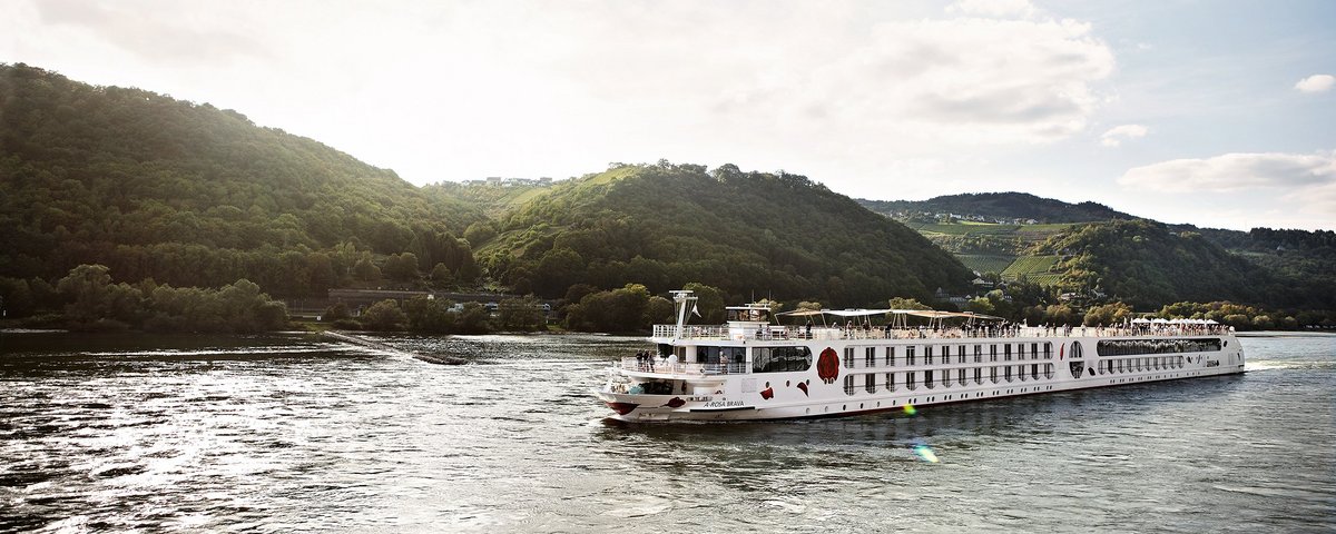 Die A-ROSA BRAVA gleitet am Nachmittag auf dem Rhein entlang, am Ufer grüne, bewaldete Hügeln und kleinen Siedlungen im Hintergrund.