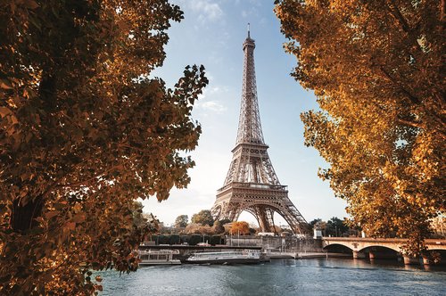Eiffelturm in Paris im Herbst, umgeben von Bäumen mit orangefarbenen Blättern, Fluss und Brücke im Vordergrund. 