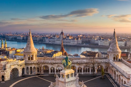 Blick auf die historische Fischerbastei in Budapest mit einer Statue eines Ritters im Vordergrund. Im Hintergrund ein Blick auf das Parlamentsgebäude von Budapest und über die Stadt bei Sonnenuntergang.