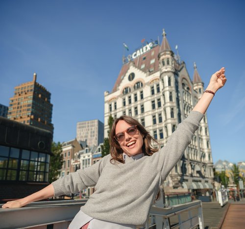 Frau mit Sonnenbrille und grauem Pullover streckt die Arme vor einem historischen Gebäude in Rotterdam aus