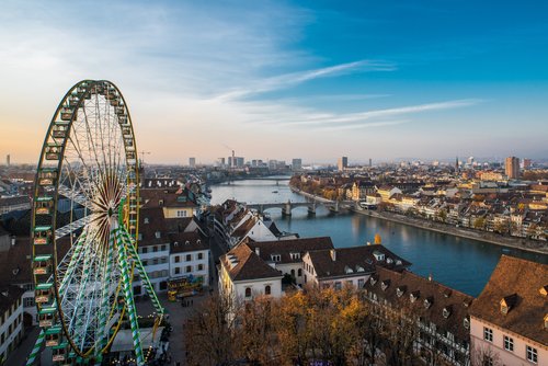 Blick auf Basel mit Rheinufer, Brücken und einem großen Riesenrad im Vordergrund bei Sonnenuntergang
