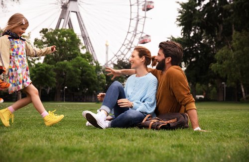 Eine Familie genießt einen sonnigen Tag im Wiener Praterpark, umgeben von grünen Bäumen. Im Hintergrund ist ein Riesenrad.