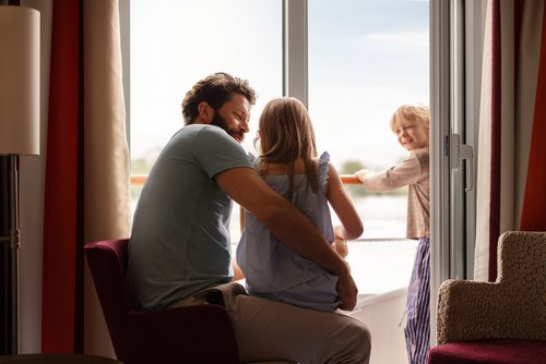 Ein Vater sitzt mit seinem Kind in einer Familienkabine auf einem Flusskreuzfahrtschiff vor dem Balkon. Ein zweites Kind steht auf dem Balkon.