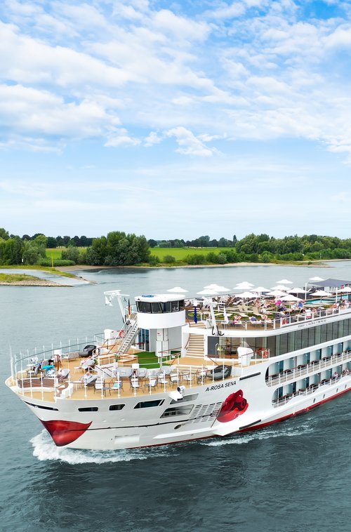 Die A-ROSA SENA fährt über den Rhein, das mit Liegestühlen und Sonnenschirmen ist gut besucht im Hintergrund das Ufer mit grüner Landschaft und Bäumen. 