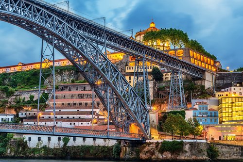 Eiserne Bogenbrücke Ponte Dom Luís I über den Fluss Douro in Porto bei Abendbeleuchtung mit Häusern und Bäumen im Hintergrund.