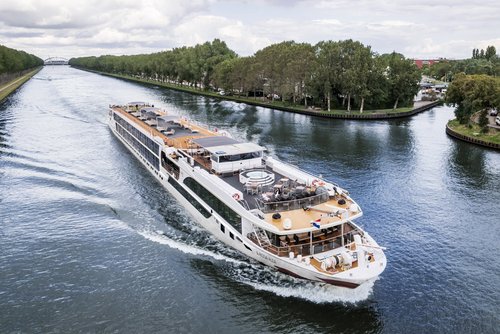Die A-ROSA ALEA fährt an einem städtischen Ufer mit Bäumen entlang, im Hintergrund eine Brücke. Das Sonnendeck ist mit Whirlpool, Sonnenliegen und Sonnensegeln ausgestattet.