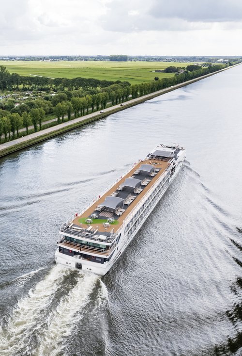 Ein Flusskreuzfahrtenschiff fährt auf dem Rhein am Ufer ein kleines Dorf, grüne Felder und Bäume. Das Sonnendeck ist mit Whirlpool, Sonnenliegen und Sonnensegeln ausgestattet.