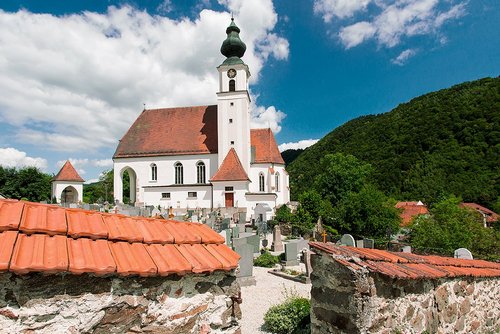 Die Pfarrkirche in Engelhartszell mit markantem Turm. Im Vordergrund ist eine Mauer und dahinter Grabsteine, im Hintergrund ein dicht bewachsener Hügel.