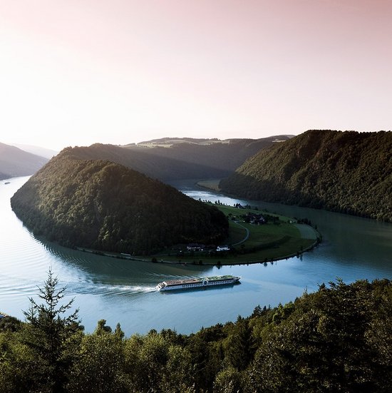 Ein Flusskreuzfahrtschiff der A-ROSA Flotte auf der Donau entlang der Schlögener Schlinge. Panoramaaufnahme mit Blick auf die angrenzende Hügellandschaft bei Sonnenaufgang.