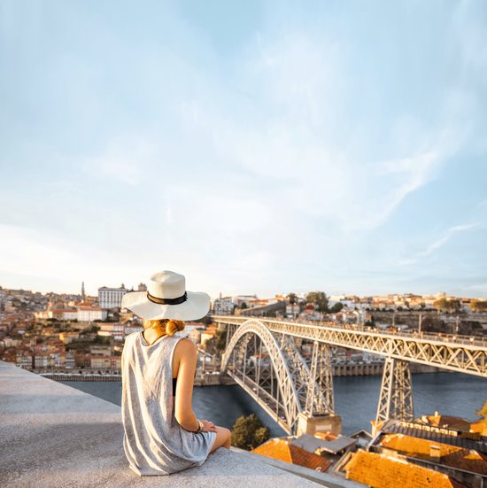 Frau mit weißem Hut und grauem Kleid sitzt auf einer Mauer und blickt auf die Stahlbogenbrücke Dom Luís I über den Fluss in Porto.