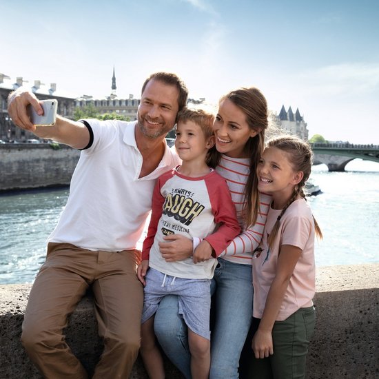 Familie mit Vater, Mutter, Sohn und Tochter macht Selfie auf Brücke über Fluss in Paris bei sonnigem Wetter.