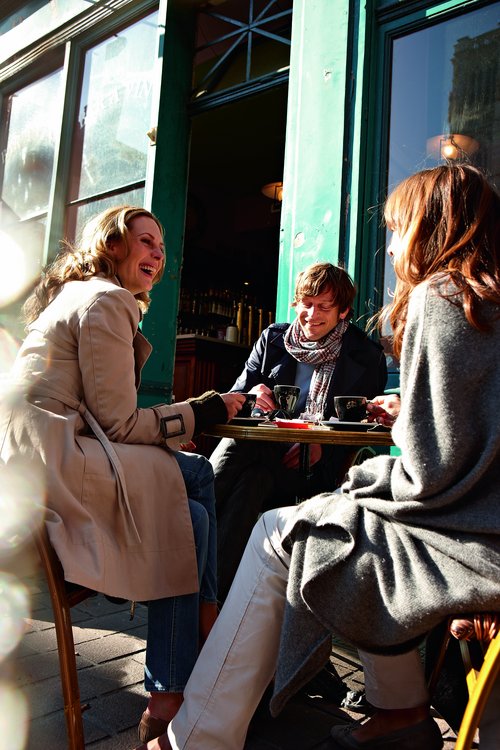 Drei Personen sitzten an einem Tisch in einem Außenbereich eines Cafés und trinken ein Heißgetränk.